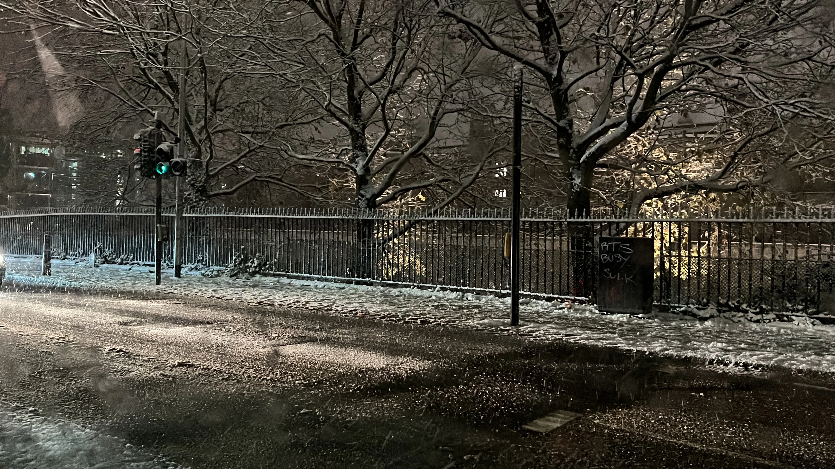 Image of a street and sidewalk lightly covered with snow. On the other side of the street is a wrought iron fence and some trees.